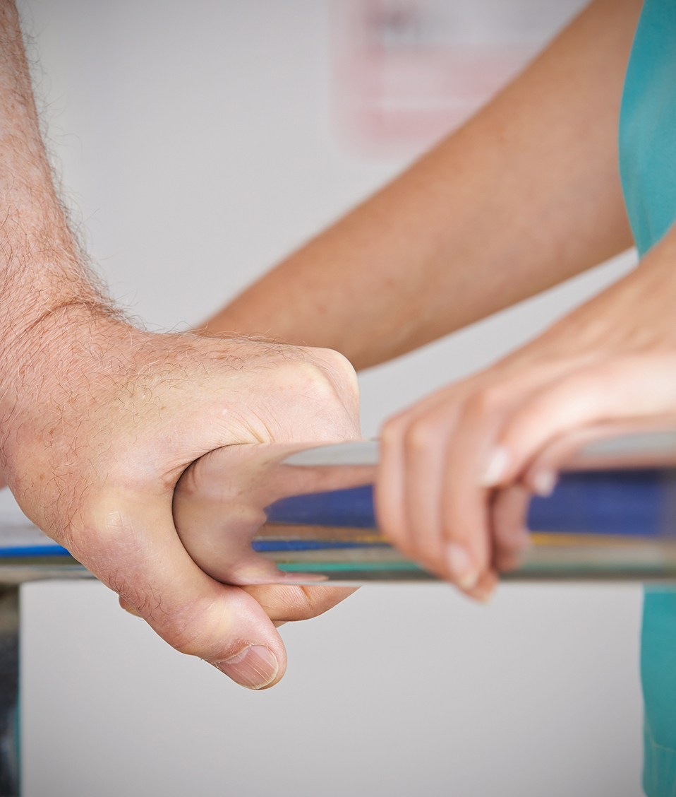 Durante un proceso de rehabilitación, la mano de una persona se sujeta a una barandilla entre los brazos de una enfermera. 